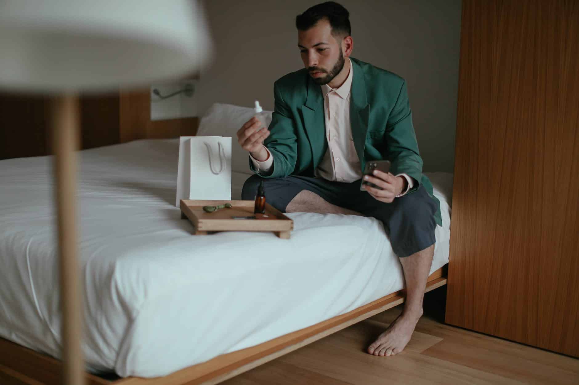 elegantly dressed man sitting on a bed and holding a cosmetic product